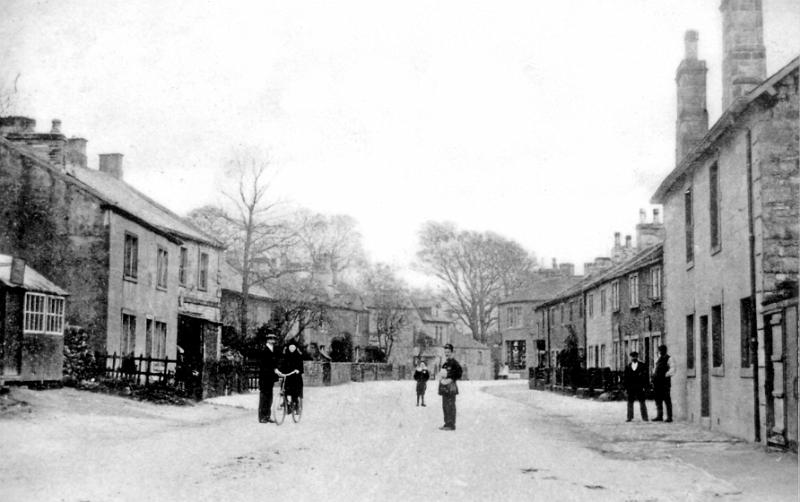 Main Street 1904.JPG - Main Street around 1904 - The Boar's Head is on the right. ( note the hut at left. ) The lady cyclist is Dorothy Wilson, Tommy Thomson, the village cobler is holding the bicycle. Bobby Stork is the boy at the back, and the postman is B. Witton. On the far right is C.Popay's greengrocers shop.